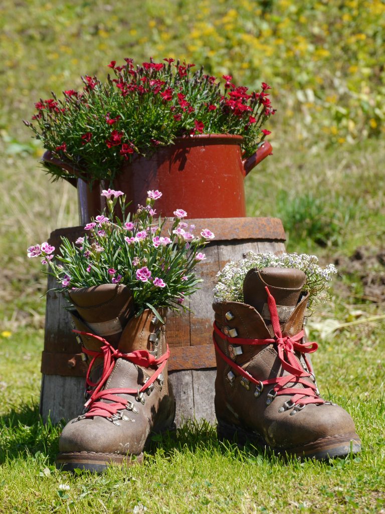 Creative floral arrangement with flowers blooming in hiking boots and red pot, set on wooden surface outdoors.